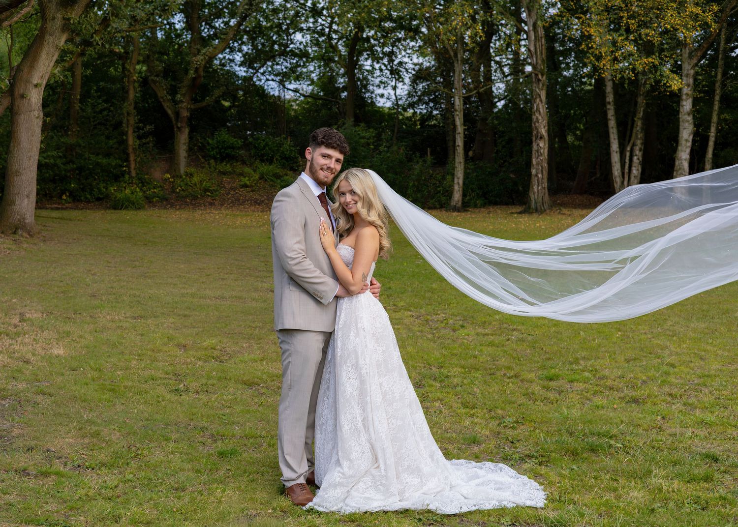 Bride and groom embracing outdoors with veil flowing in wind, surrounded by lush green trees on their wedding day.