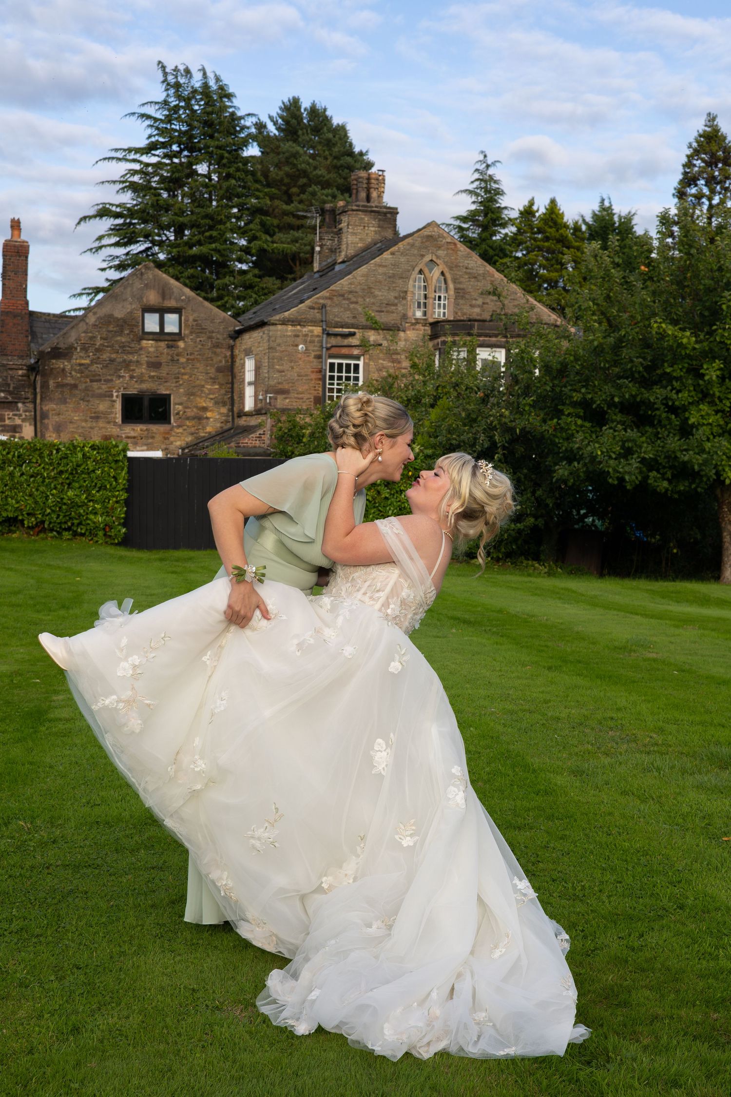 Two brides share a romantic dip kiss on green lawn outside a stone manor house on their wedding day.