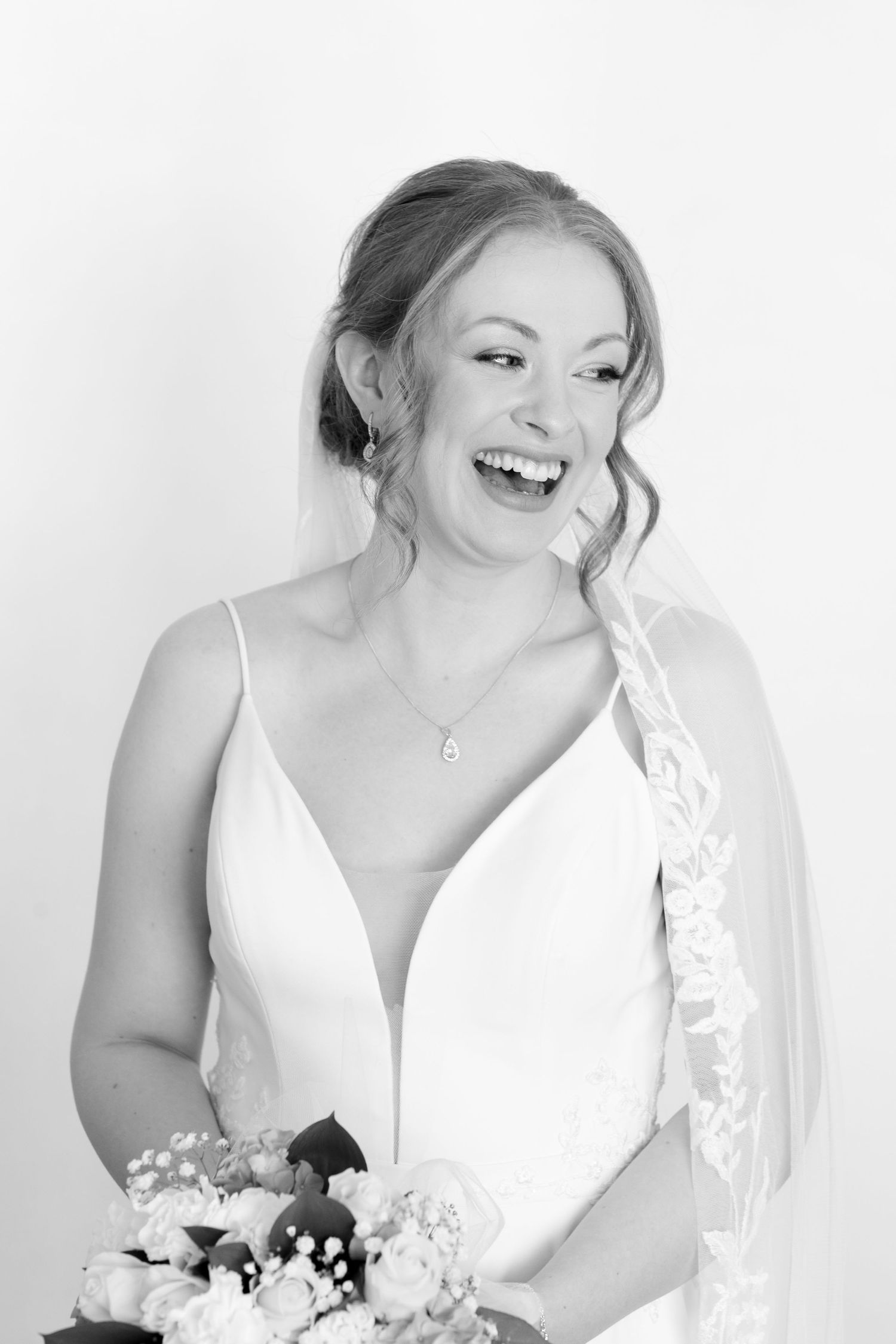 Joyful bride in white v-neck wedding dress with veil laughs in black and white portrait.