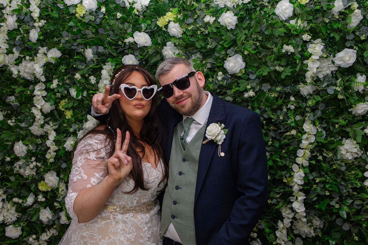 A playful wedding moment against a wall of white flowers and greenery.