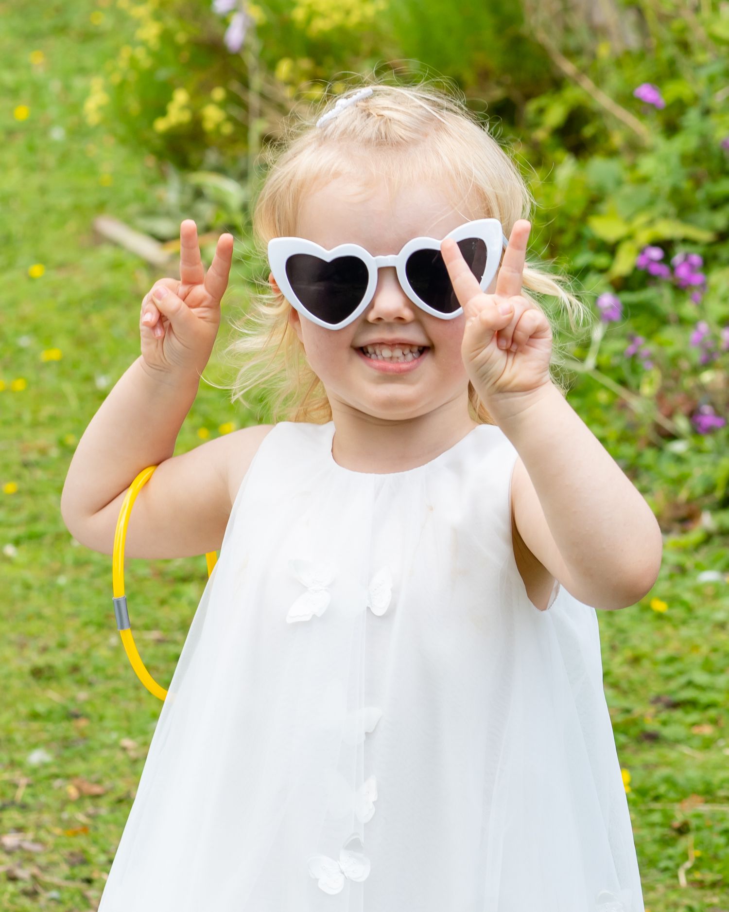 A small child wearing heart-shaped white sunglasses and a white dress makes peace signs while smiling in a garden setting.