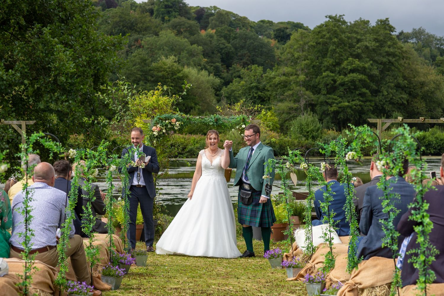 Wedding ceremony guests gather outdoors in a lush garden setting surrounded by blooming flowers and greenery.