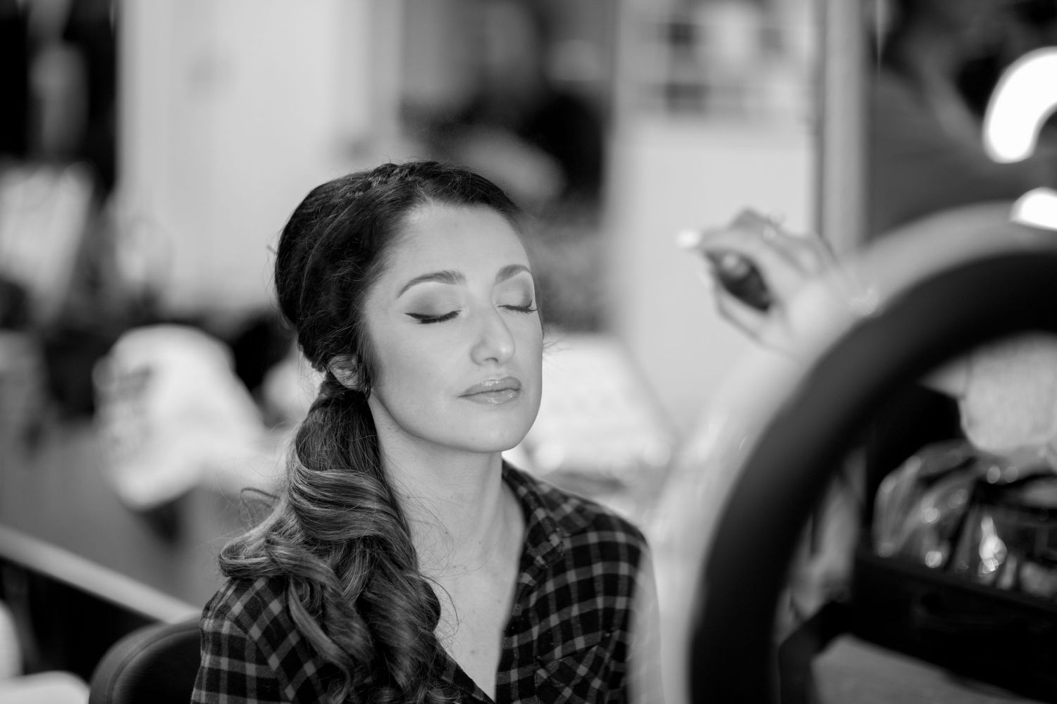 A person in a plaid shirt closes their eyes while getting makeup applied by a professional makeup artist.