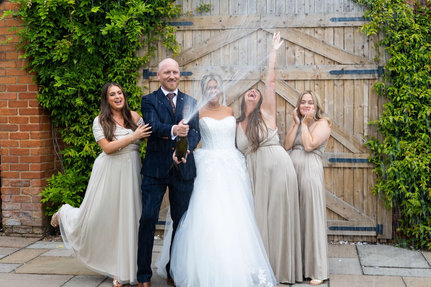 Bride in white gown pops champagne with groom and bridesmaids in taupe dresses outside rustic barn doors.