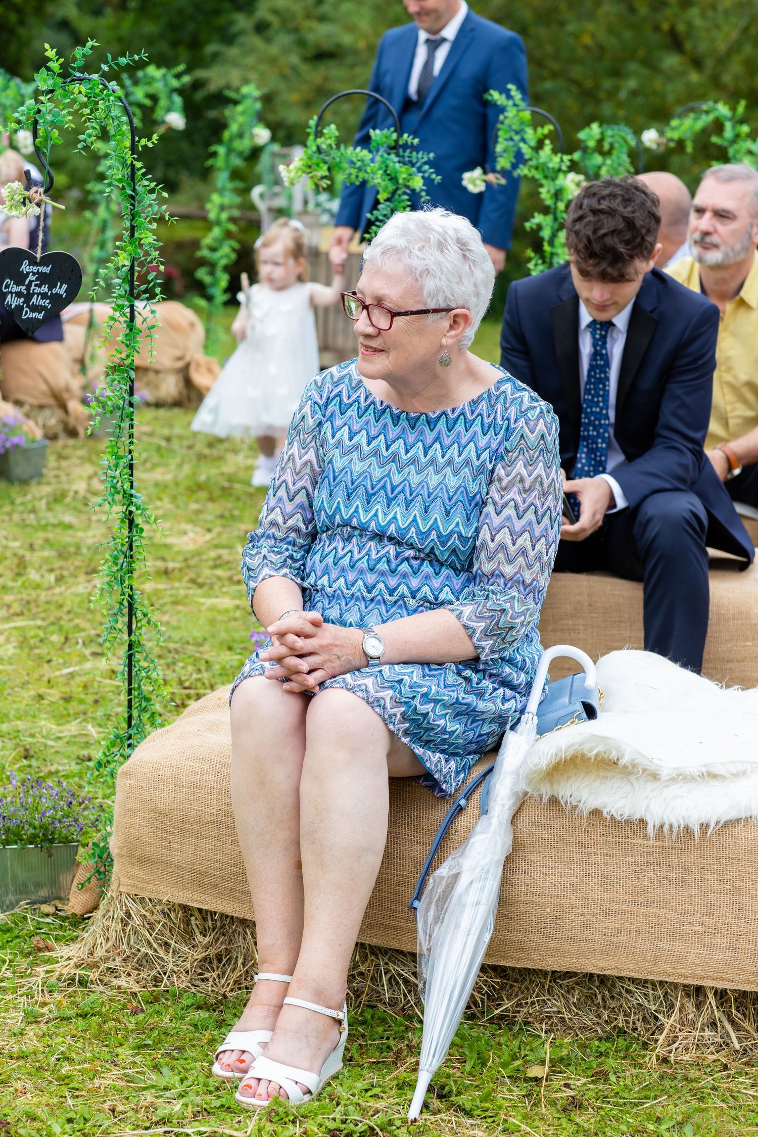 A person in a blue patterned dress sits on a rustic hay bale seating arrangement at an outdoor event with a white umbrella.