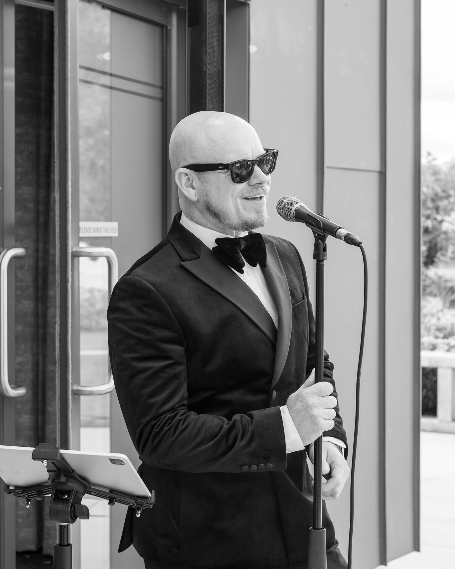 Three men in black suits and sunglasses stand outside glass doors in a formal security or professional setting.