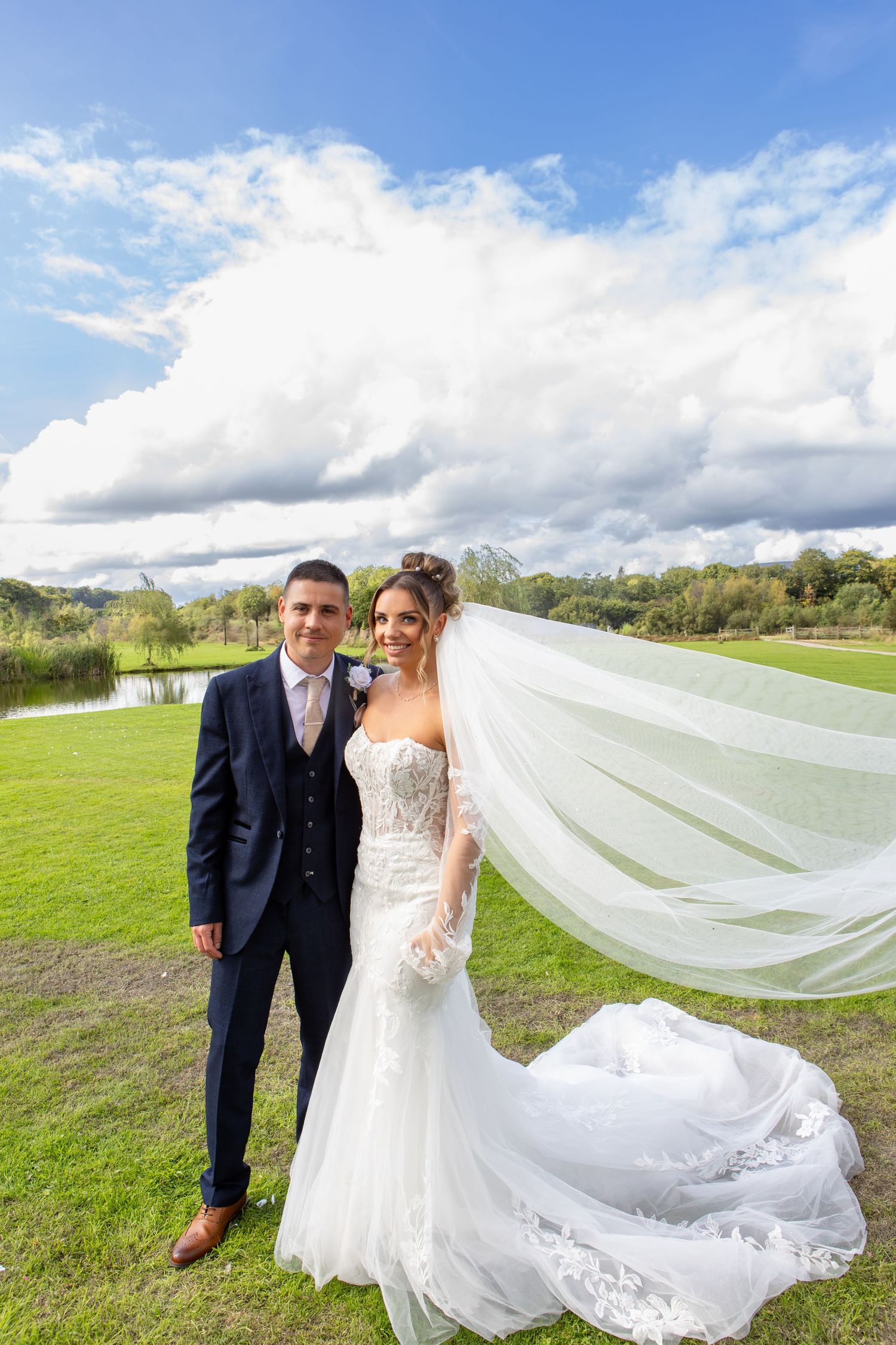 A couple poses outdoors on their wedding day with a flowing veil against a scenic landscape with clouds and greenery.