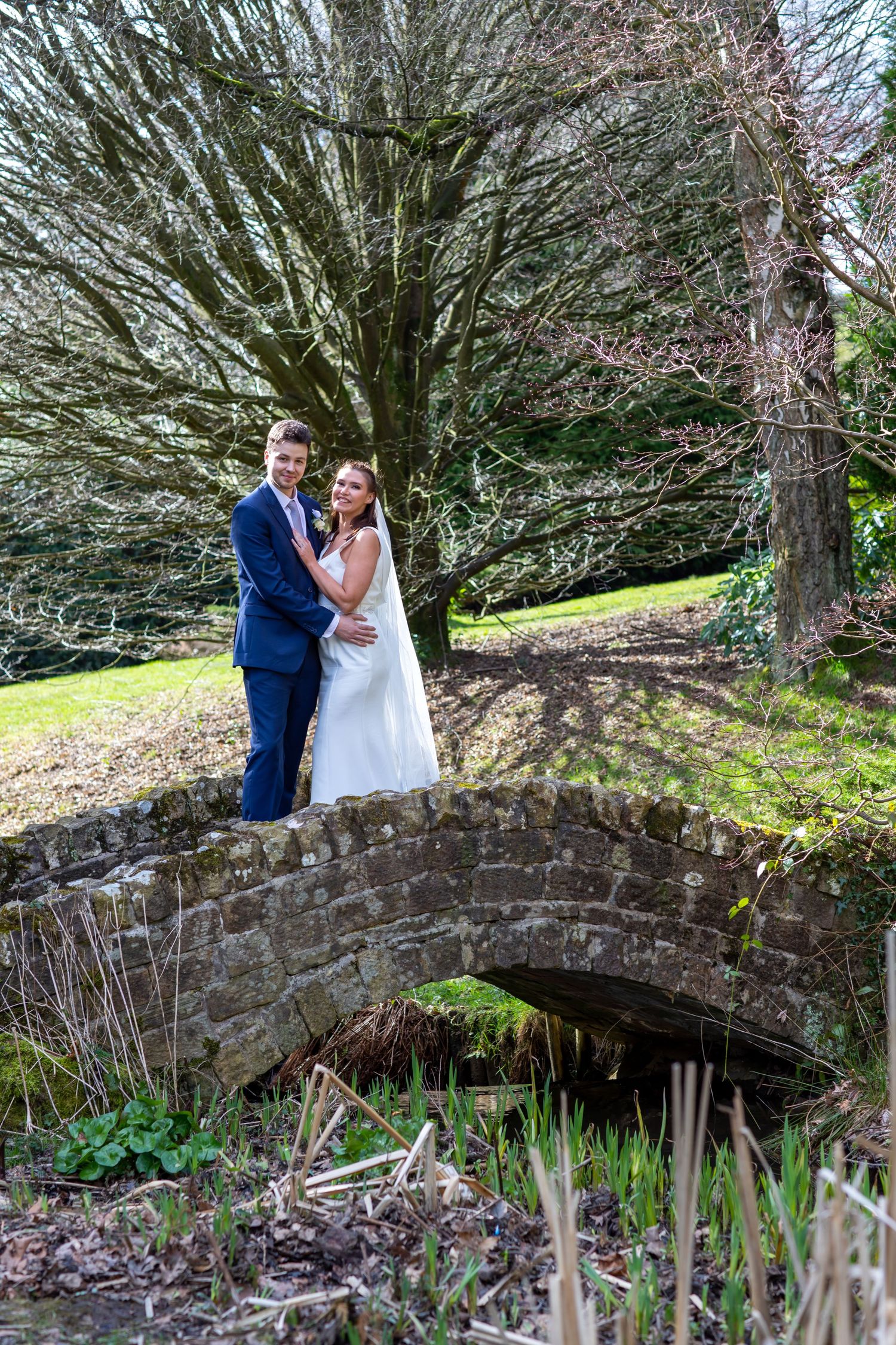 A wedding couple stands on a rustic stone bridge surrounded by winter trees.