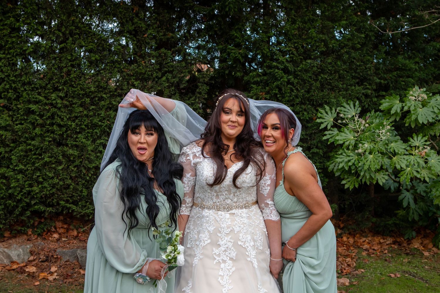 Wedding party members in sage green dresses gather outdoors for group photos in a lush garden setting.