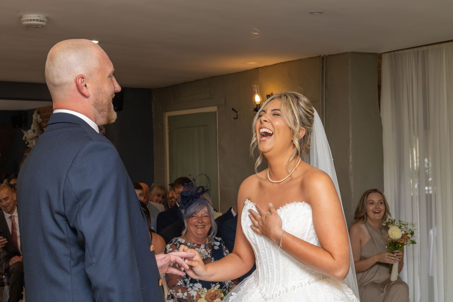 Bride laughing joyfully with groom during wedding ceremony, guests smiling in background.
