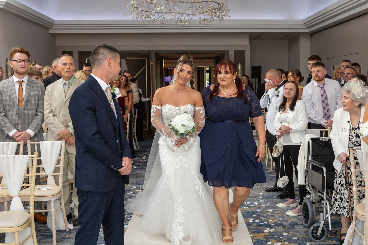 Wedding ceremony at an elegant indoor venue with guests seated on white chairs and scattered petals on the floor.