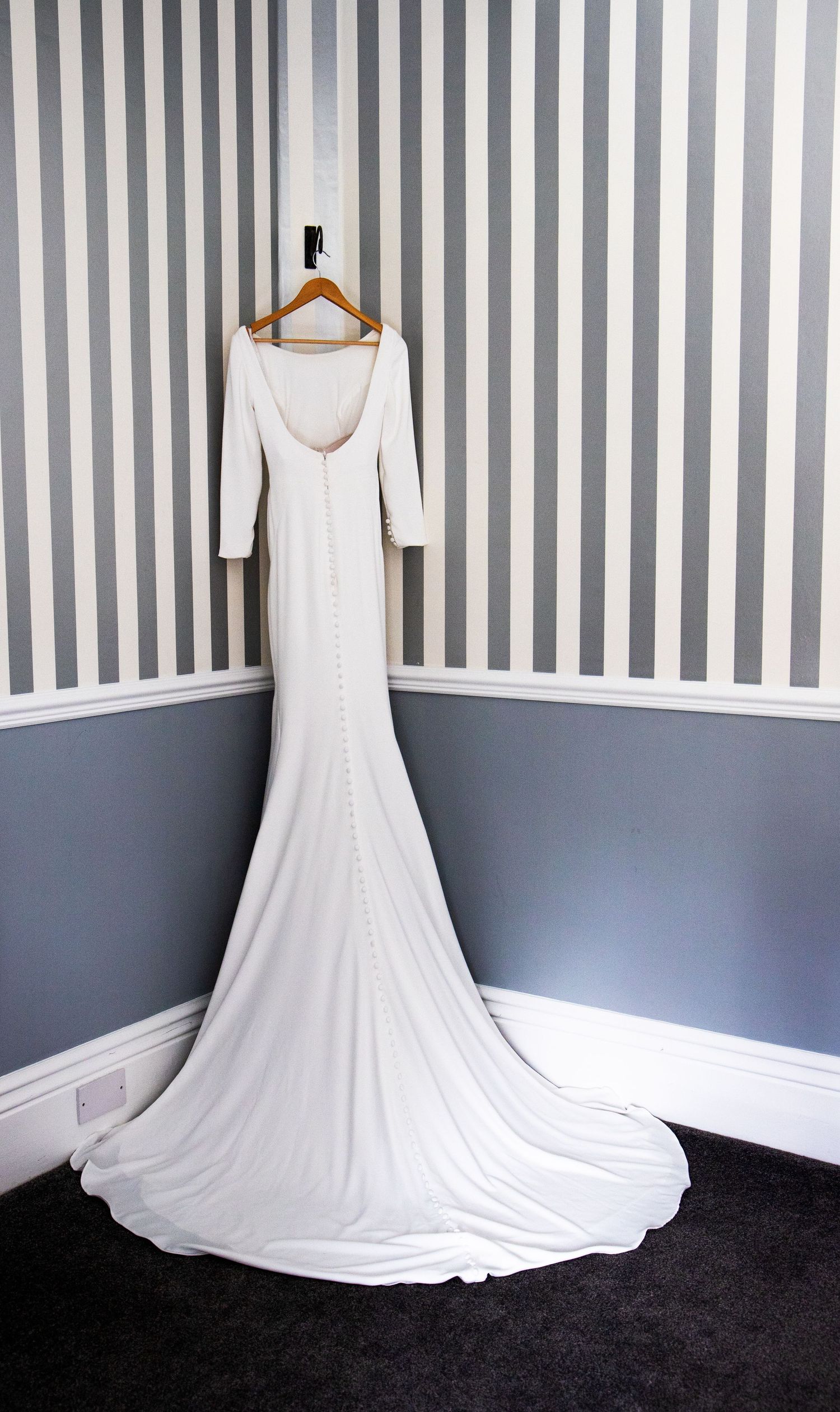 Elegant white wedding gown with long train displayed on hanger against striped wall.