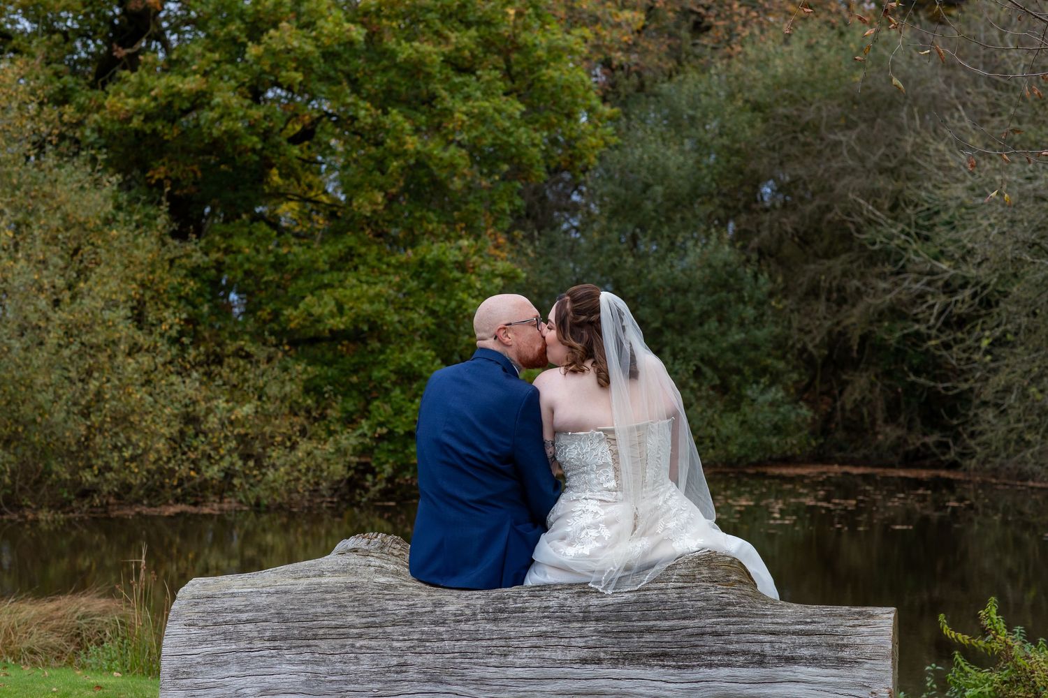 A couple in wedding attire sits together on a rustic wooden bench overlooking a tranquil pond surrounded by autumn trees.