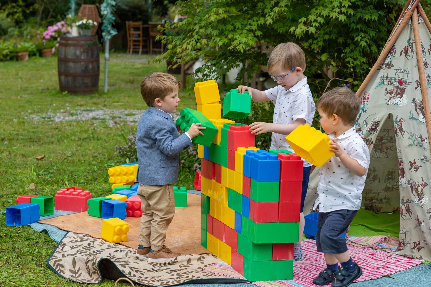 Children playing together with colorful building blocks and toys on a blanket in an outdoor garden setting.