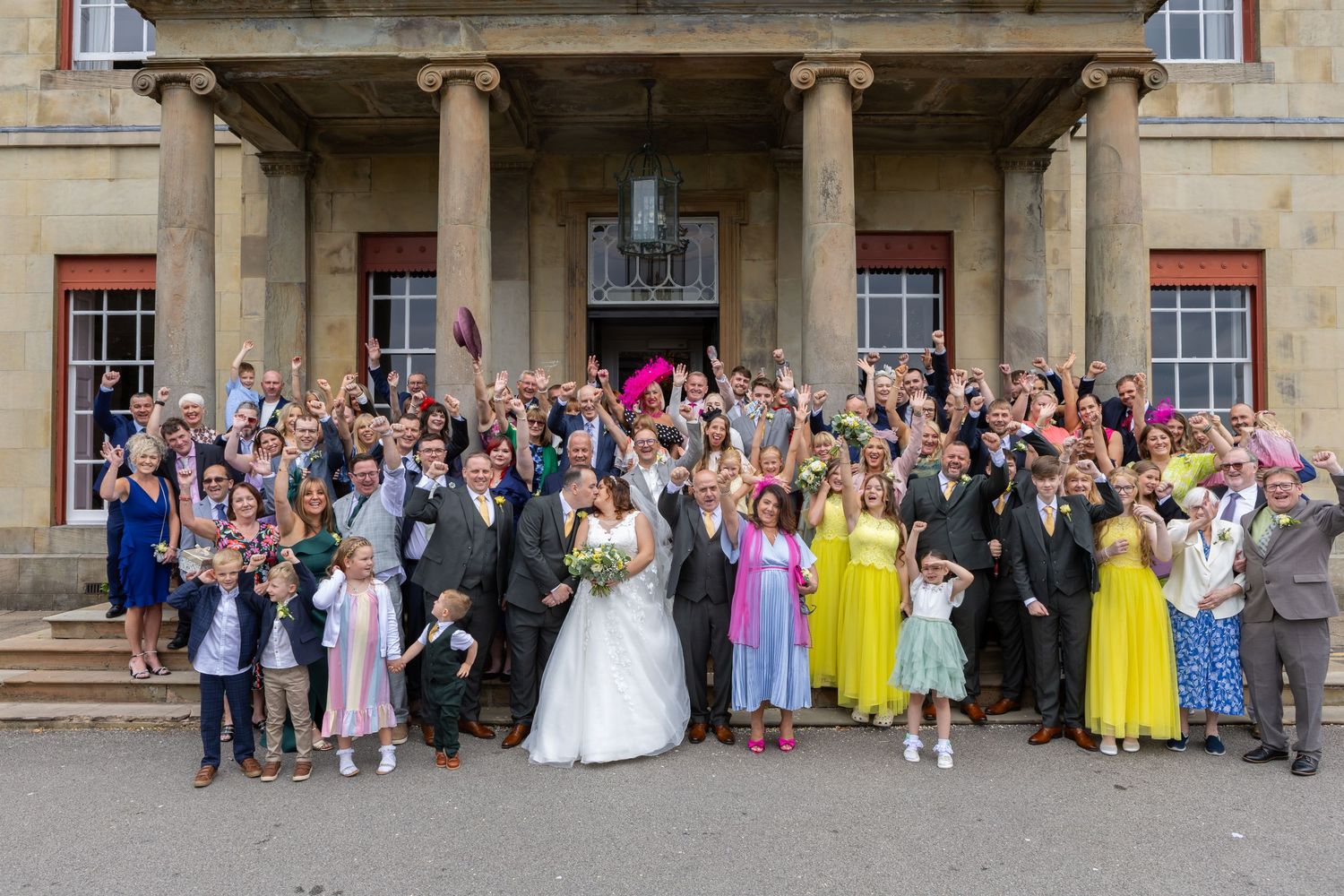 Wedding party and guests gathered for formal group photo outside historic building.