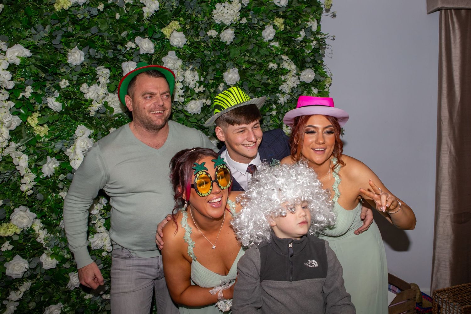 A group of people wearing colorful party hats and accessories pose together in a photo booth with greenery backdrop.