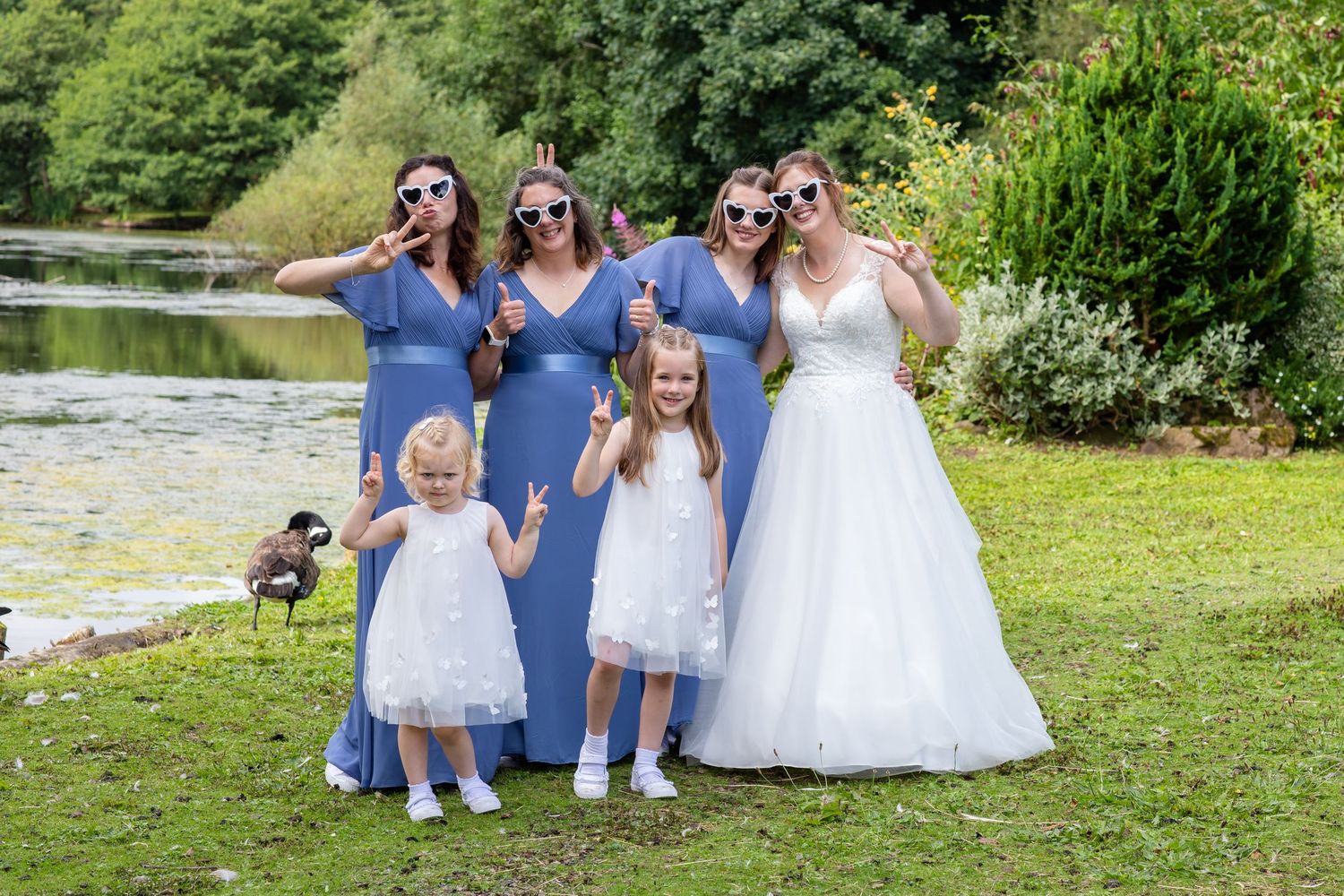 Wedding party taking group photos outdoors on grassy lawn near lake.