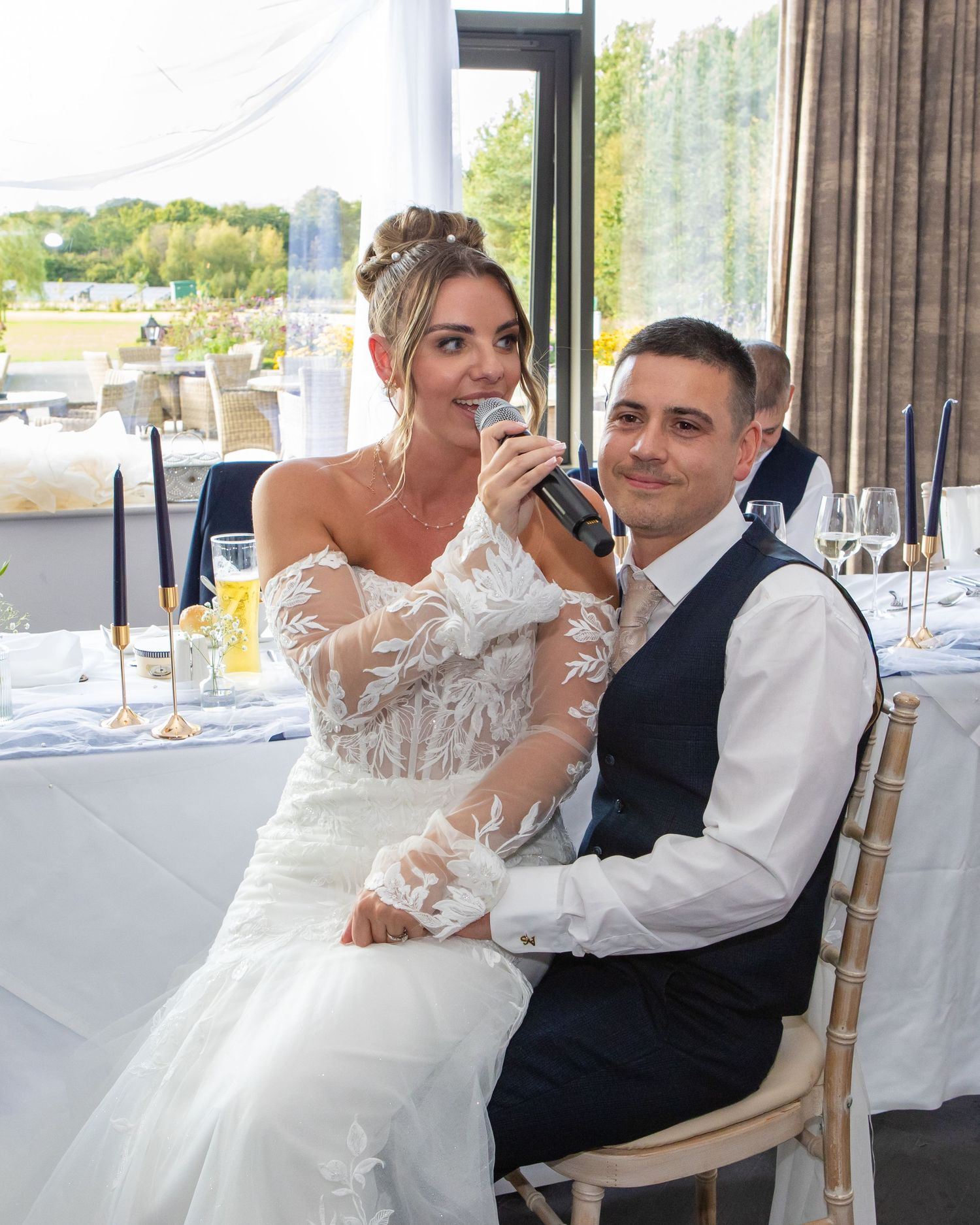 A couple at their wedding reception seated at a decorated table with the bride holding a microphone during speeches.