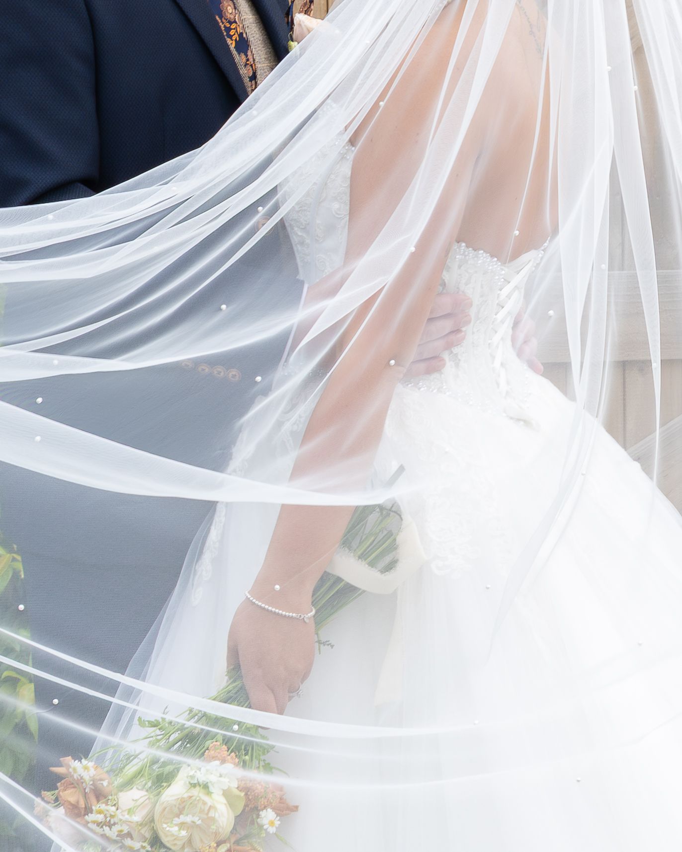 Bride in white lace gown holding bouquet, veil billowing around couple in romantic wedding photo.