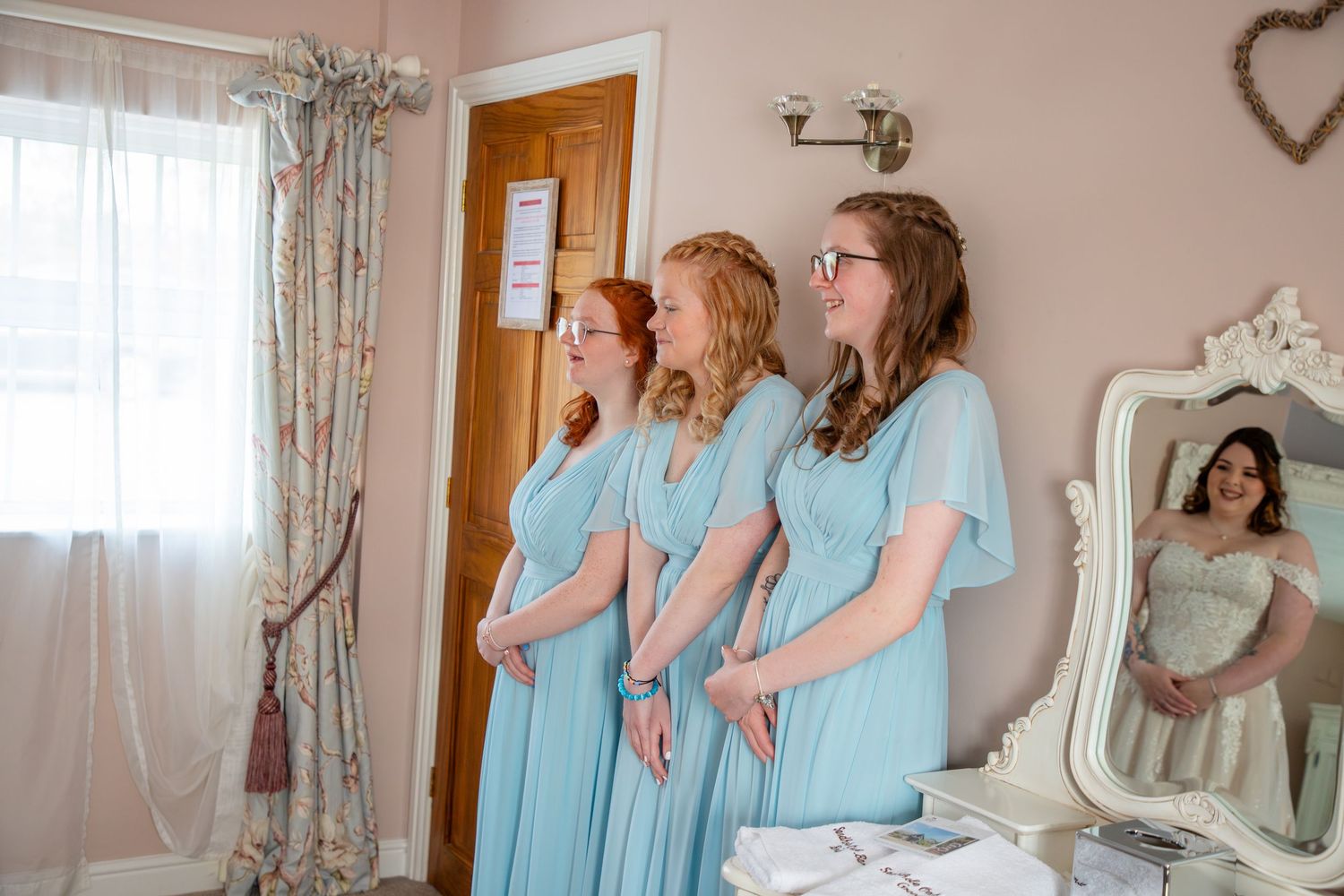 A group wearing matching light blue dresses stands in a row by a window in a bridal preparation room.