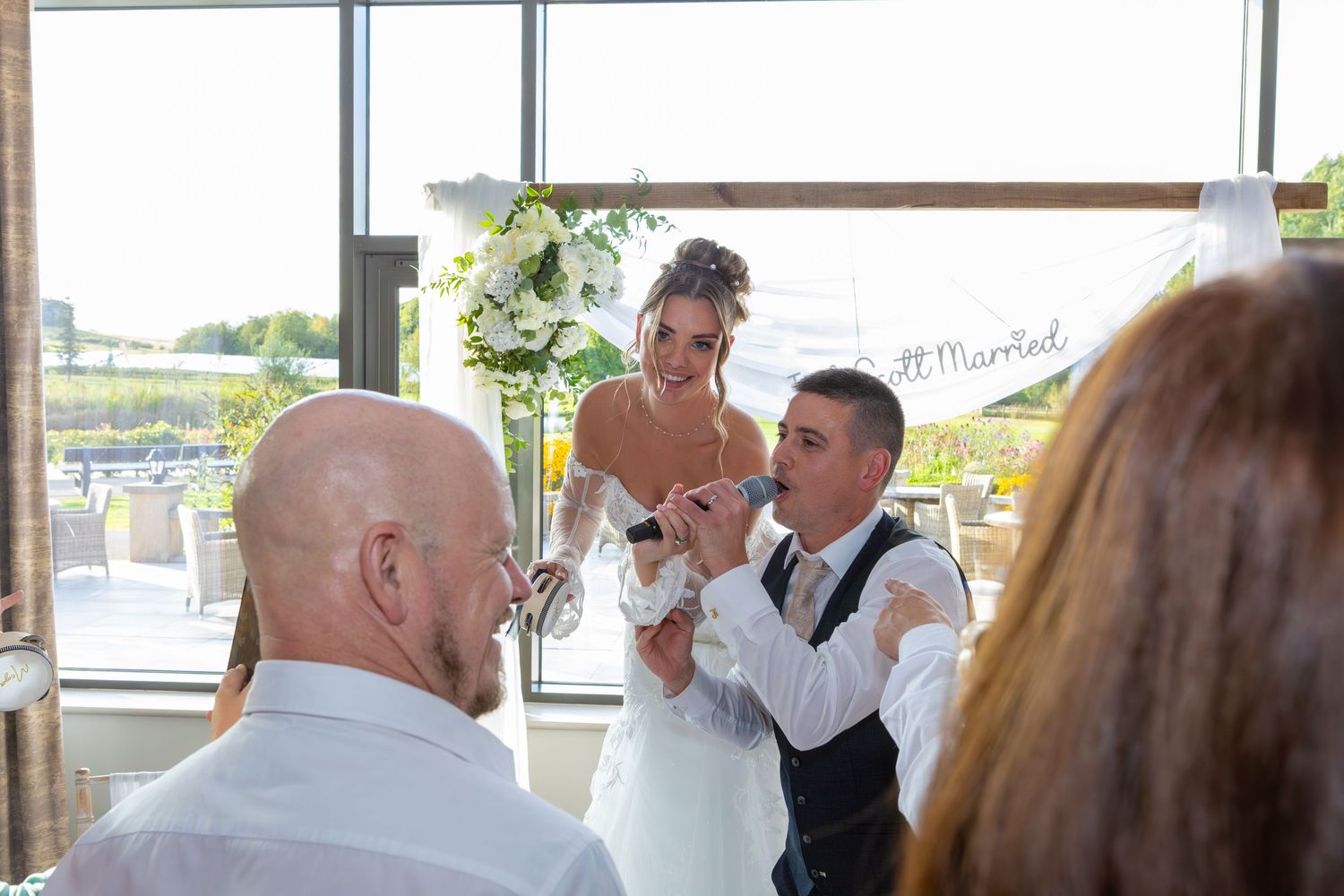 A wedding celebration moment captured at a venue with glass windows and natural lighting in the background.