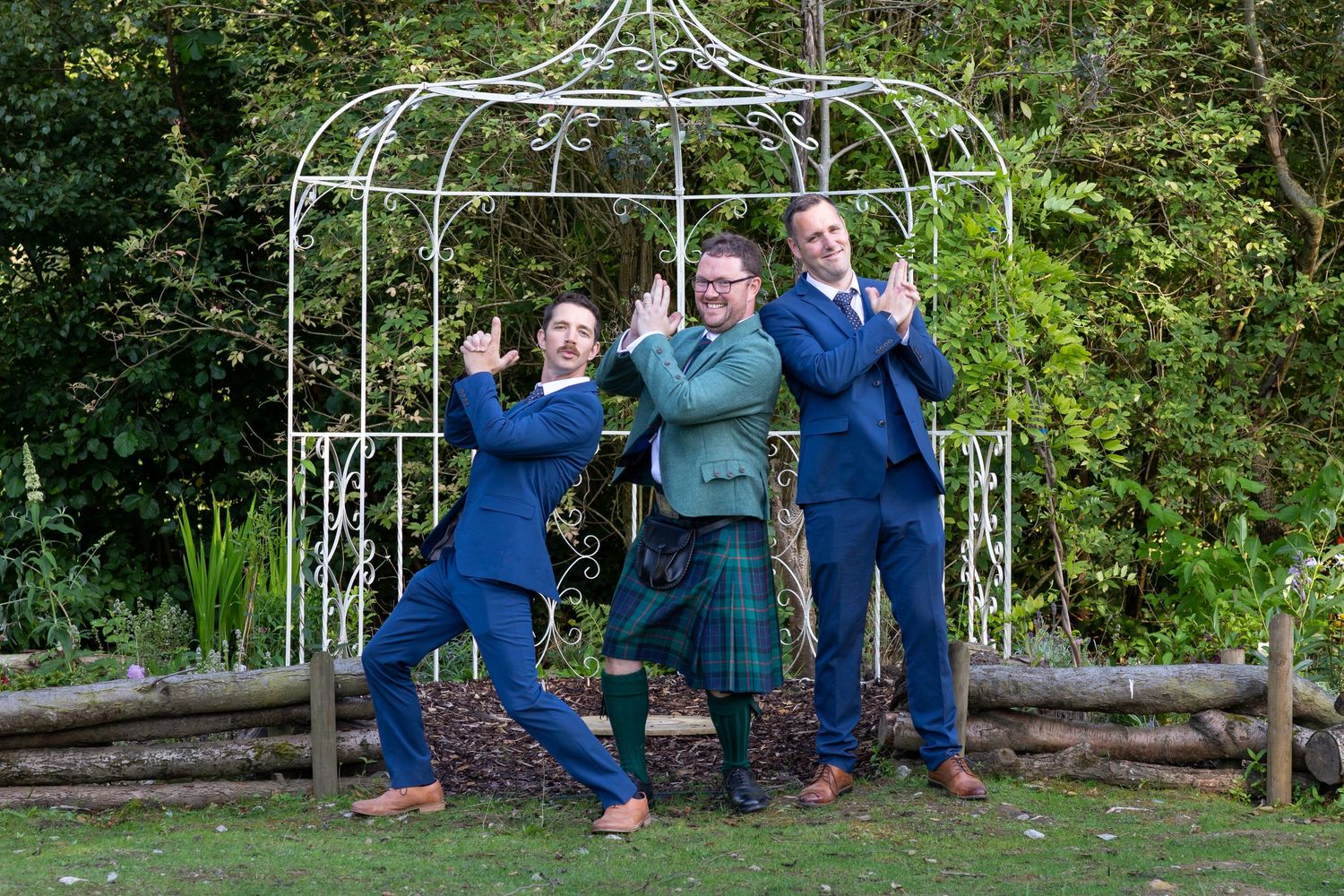 Three people strike playful poses under white garden arch outdoors.