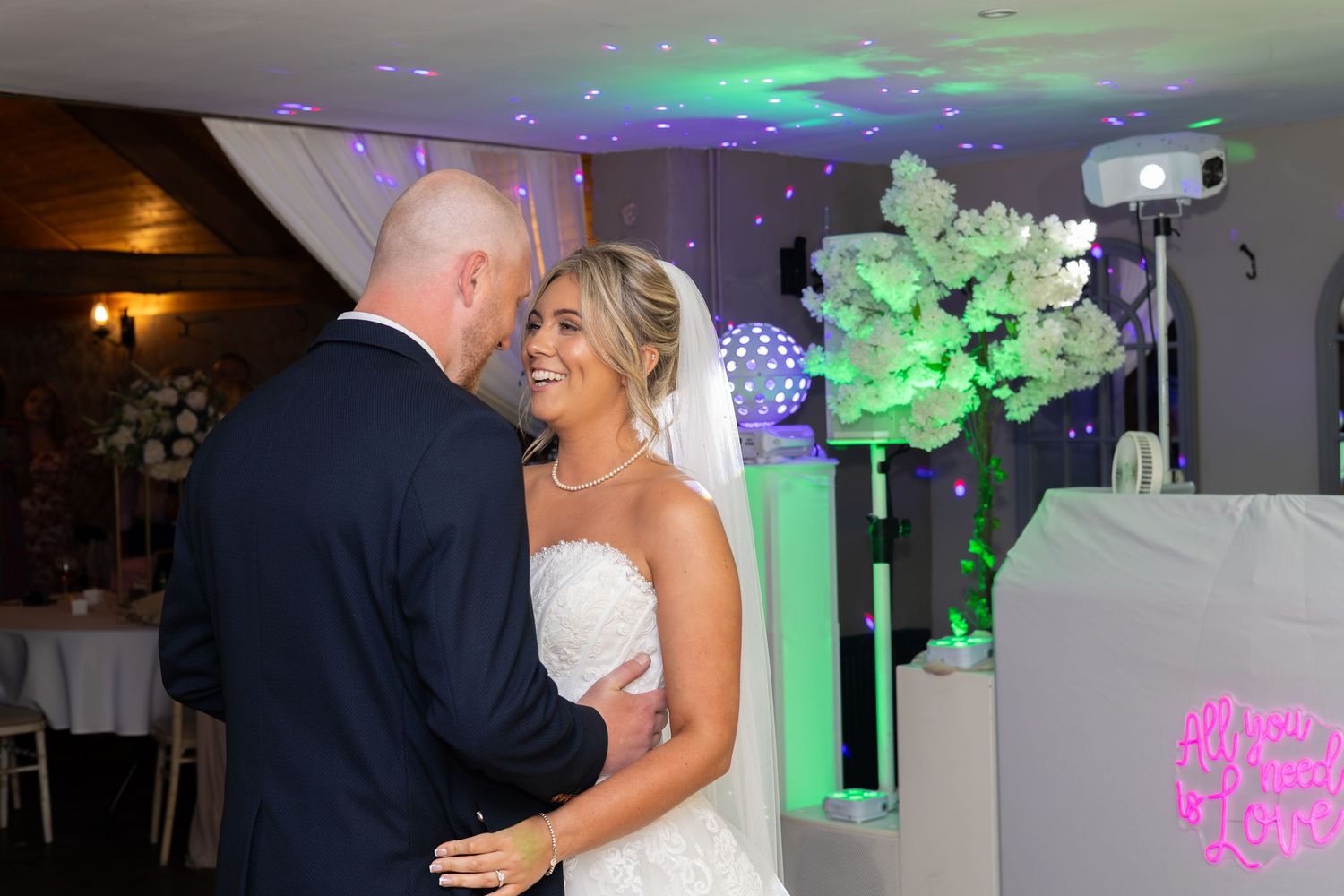 Bride and groom share first dance at wedding reception with colorful DJ lights and floral decorations in background.