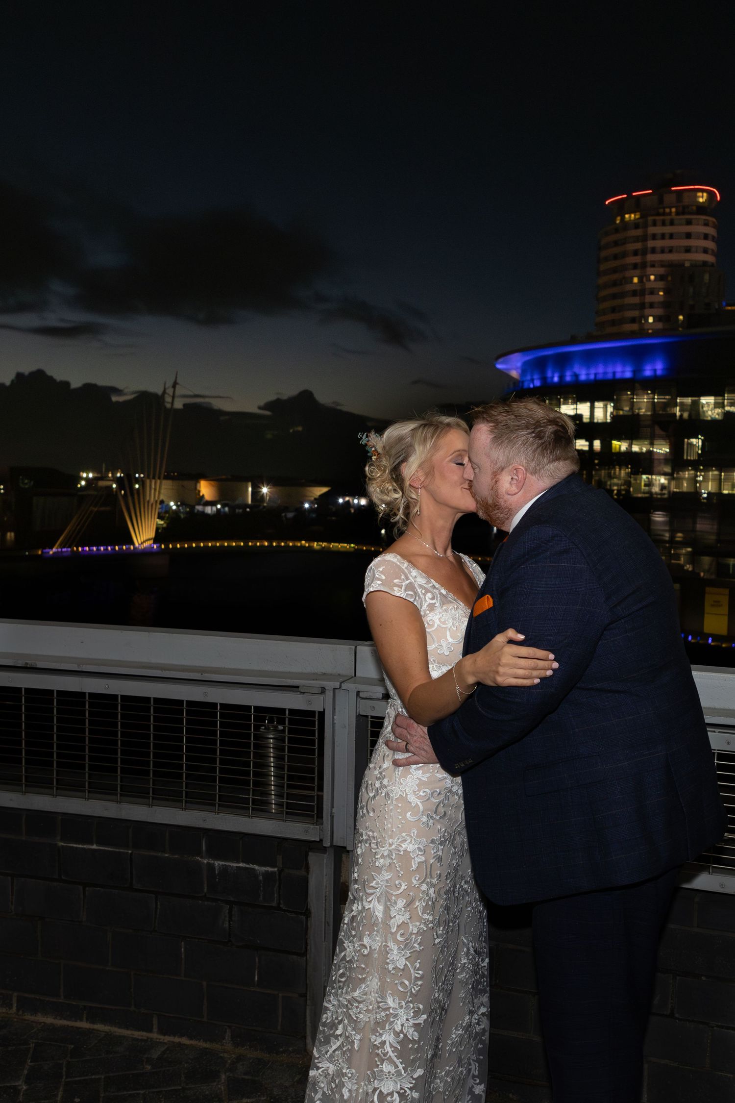 A couple shares an intimate moment at dusk on a waterfront terrace with city lights glowing in the background.