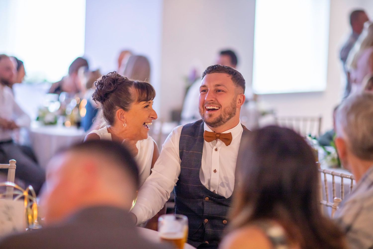 A group of people enjoying themselves at a wedding reception with laughter and smiles around dining tables.
