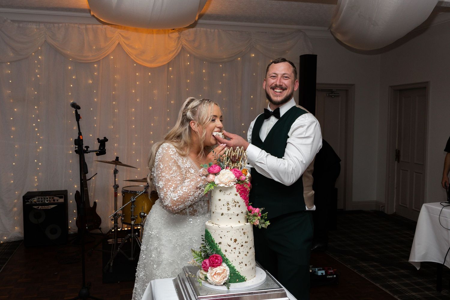 Bride and groom laughing while cutting their white wedding cake decorated with pink flowers at their reception.