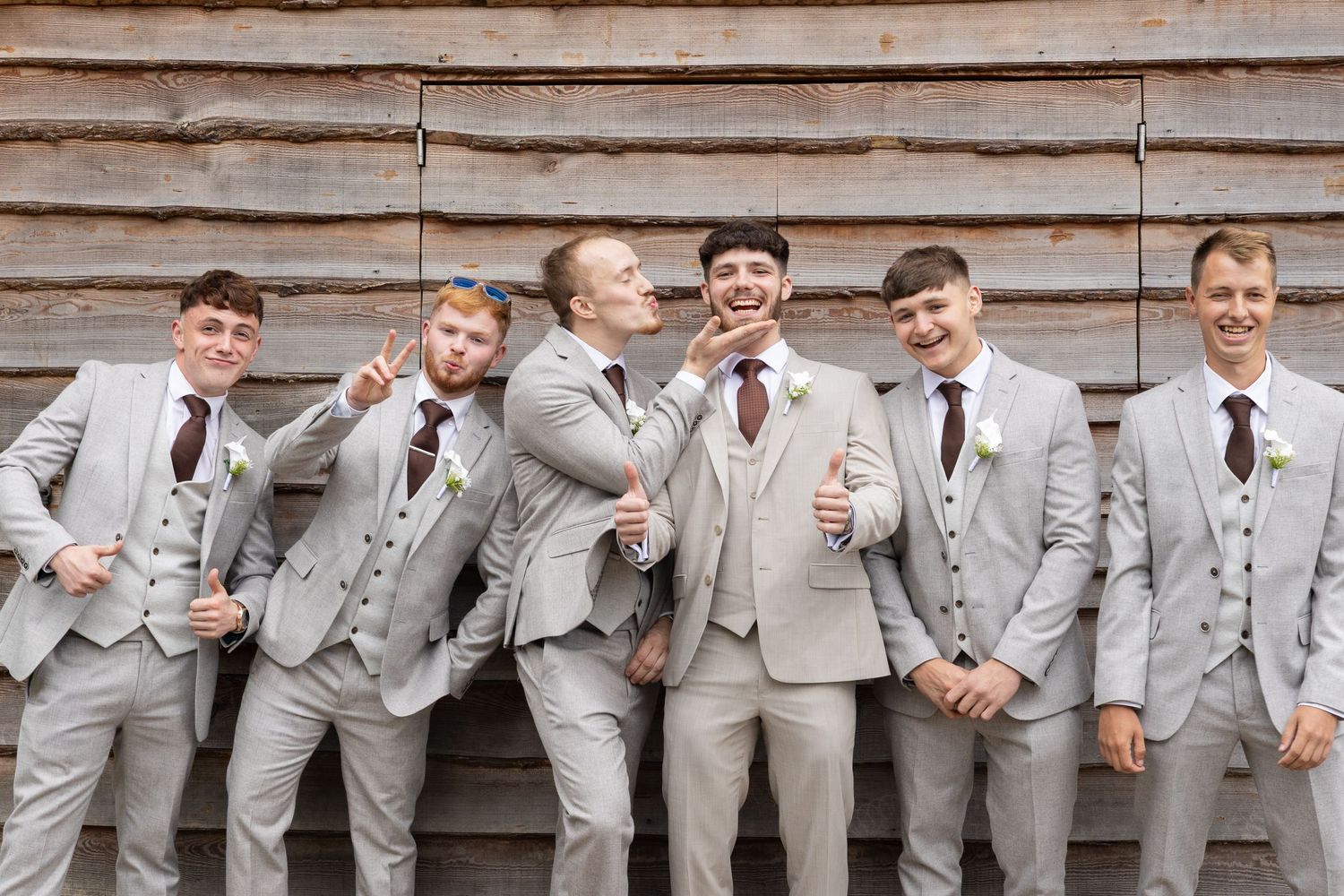Six groomsmen in matching light gray suits with burgundy ties pose playfully against a rustic wooden barn wall.