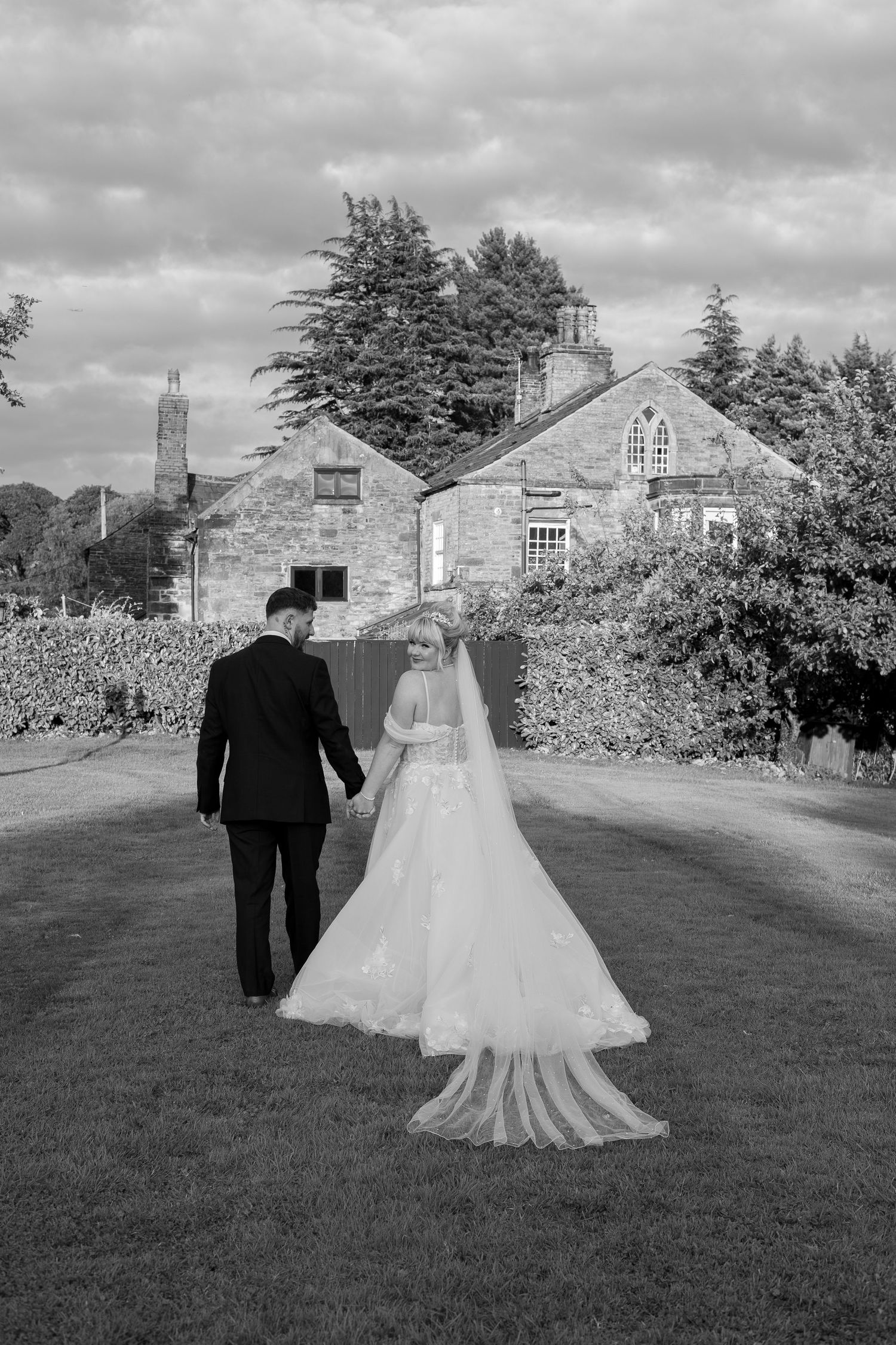 Black and white photo of a bride and groom walking hand in hand toward a stone manor house on their wedding day.