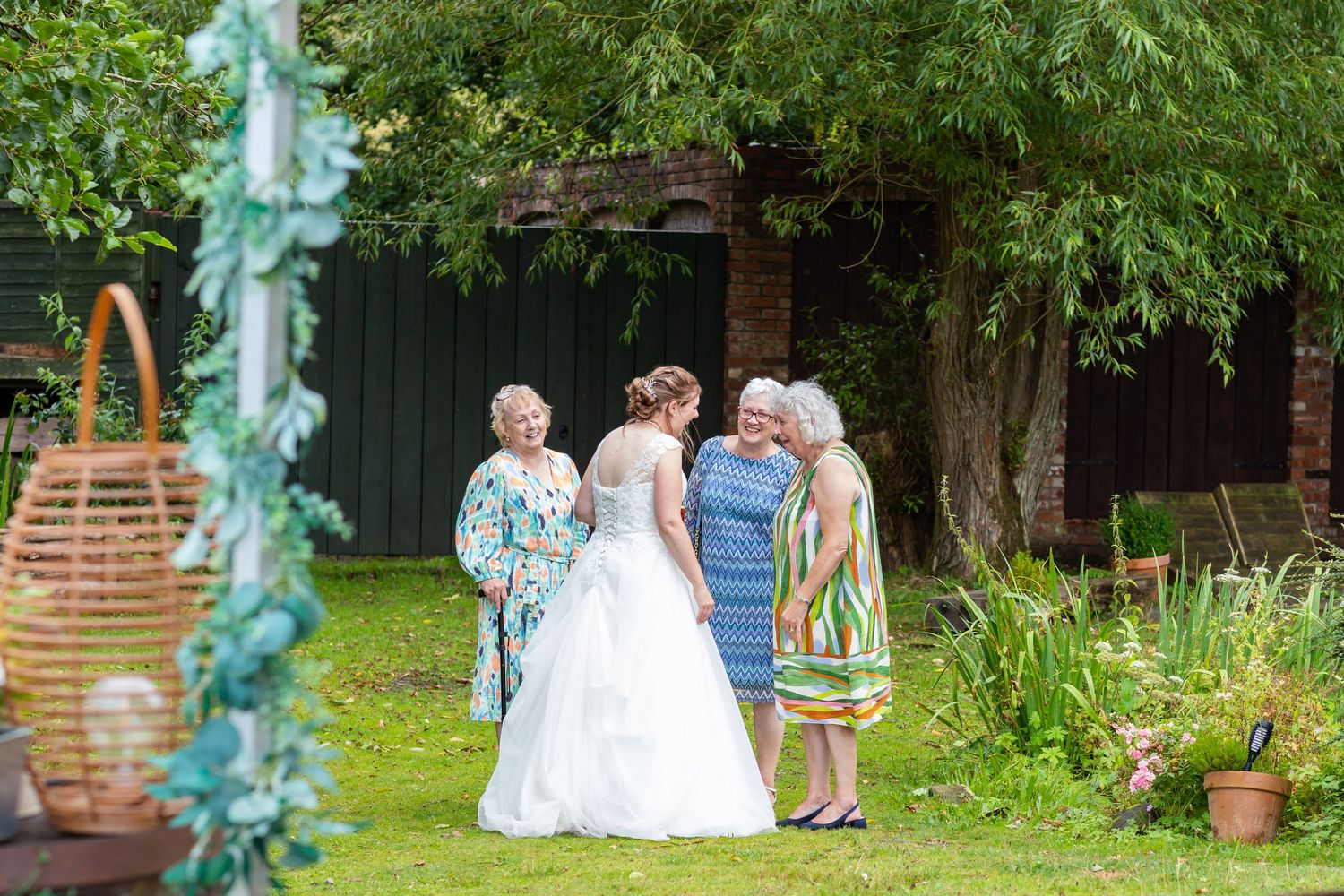 A wedding celebration takes place in a lush garden setting with decorative blue-green ribbons hanging from trees.