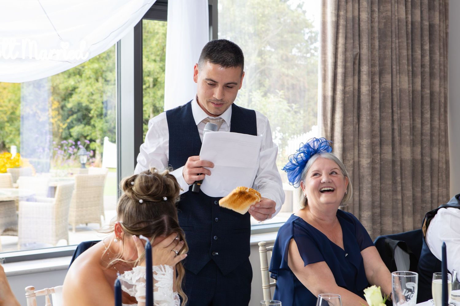Wedding guests enjoy a meal at elegantly decorated tables in a bright reception venue with large windows.