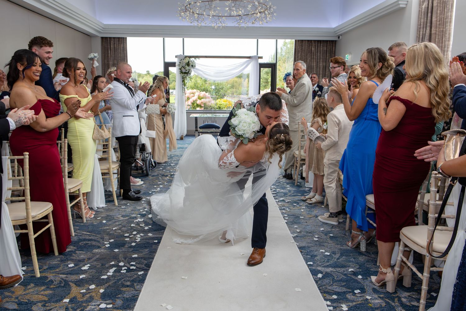 Wedding guests dance and celebrate together in an elegant ballroom with a chandelier and large windows.