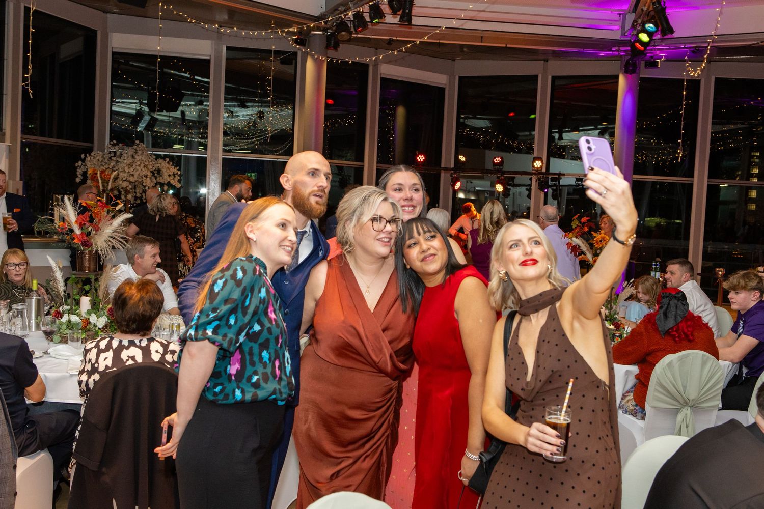 People dancing and celebrating at an indoor event venue with large windows and purple mood lighting.