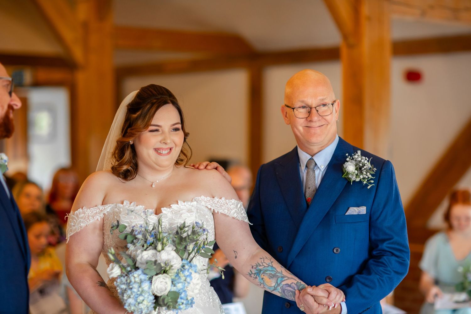 A bride wearing an off-shoulder white gown walks down the aisle at a rustic wooden beam venue with floral decorations.