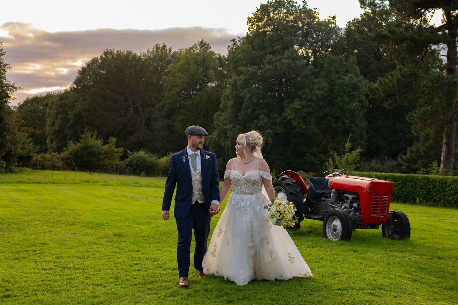 Bride and groom walk on lush green lawn at sunset beside a vintage red tractor at an outdoor wedding venue.