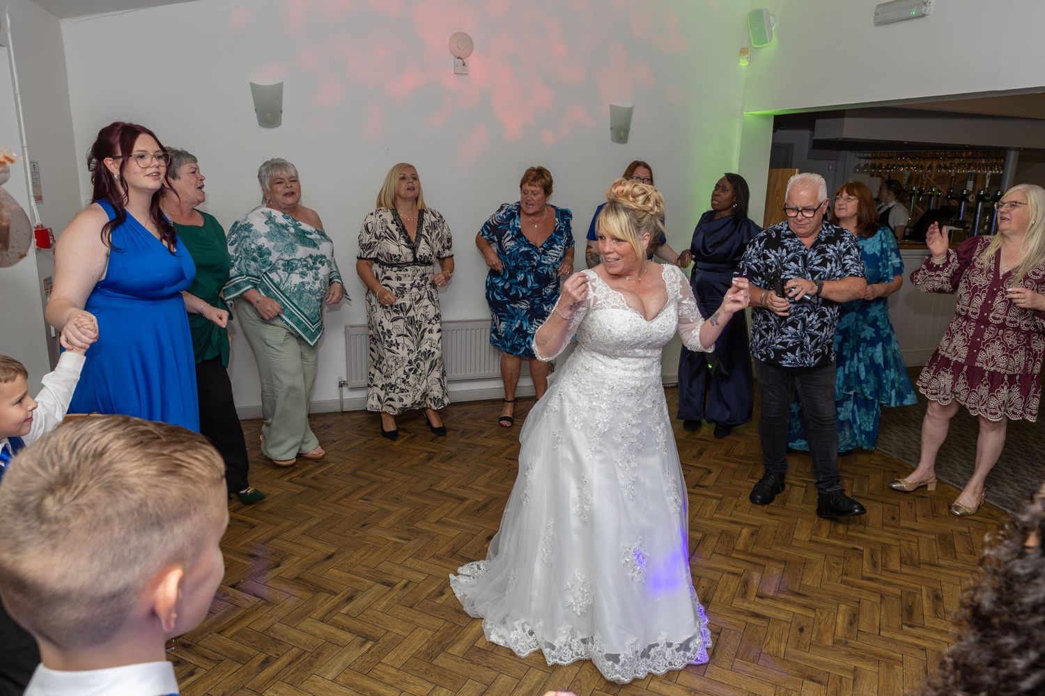 Bride in white wedding gown dancing joyfully with guests at a wedding reception on a wooden dance floor.