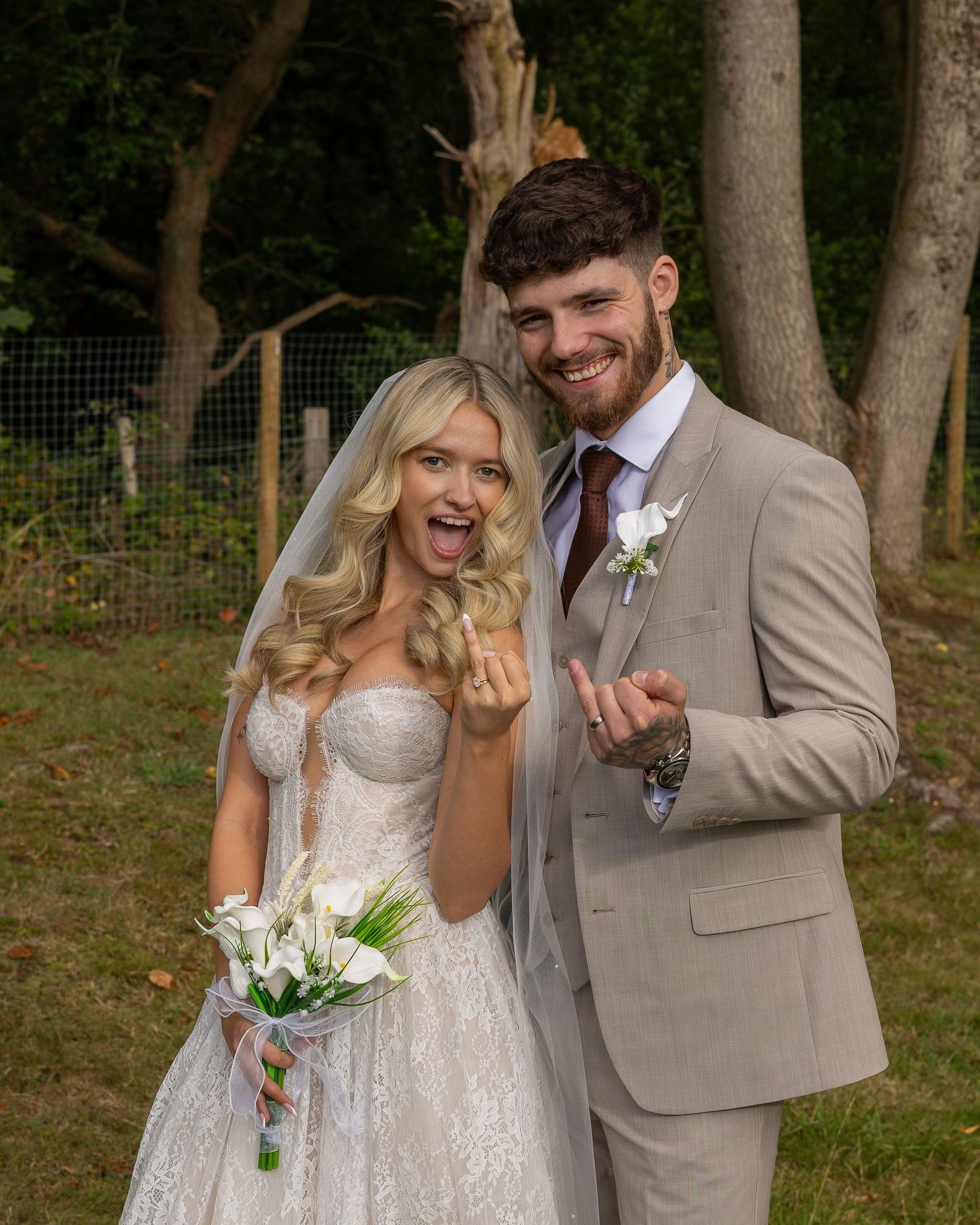 A smiling bride and groom in wedding attire show off their rings outdoors, with the bride holding white flowers.