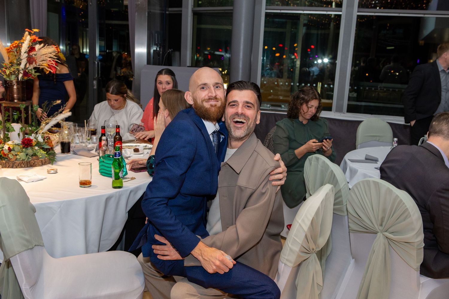 Two people in formal attire smile together at a wedding reception with white tablecloths and decorative table settings.