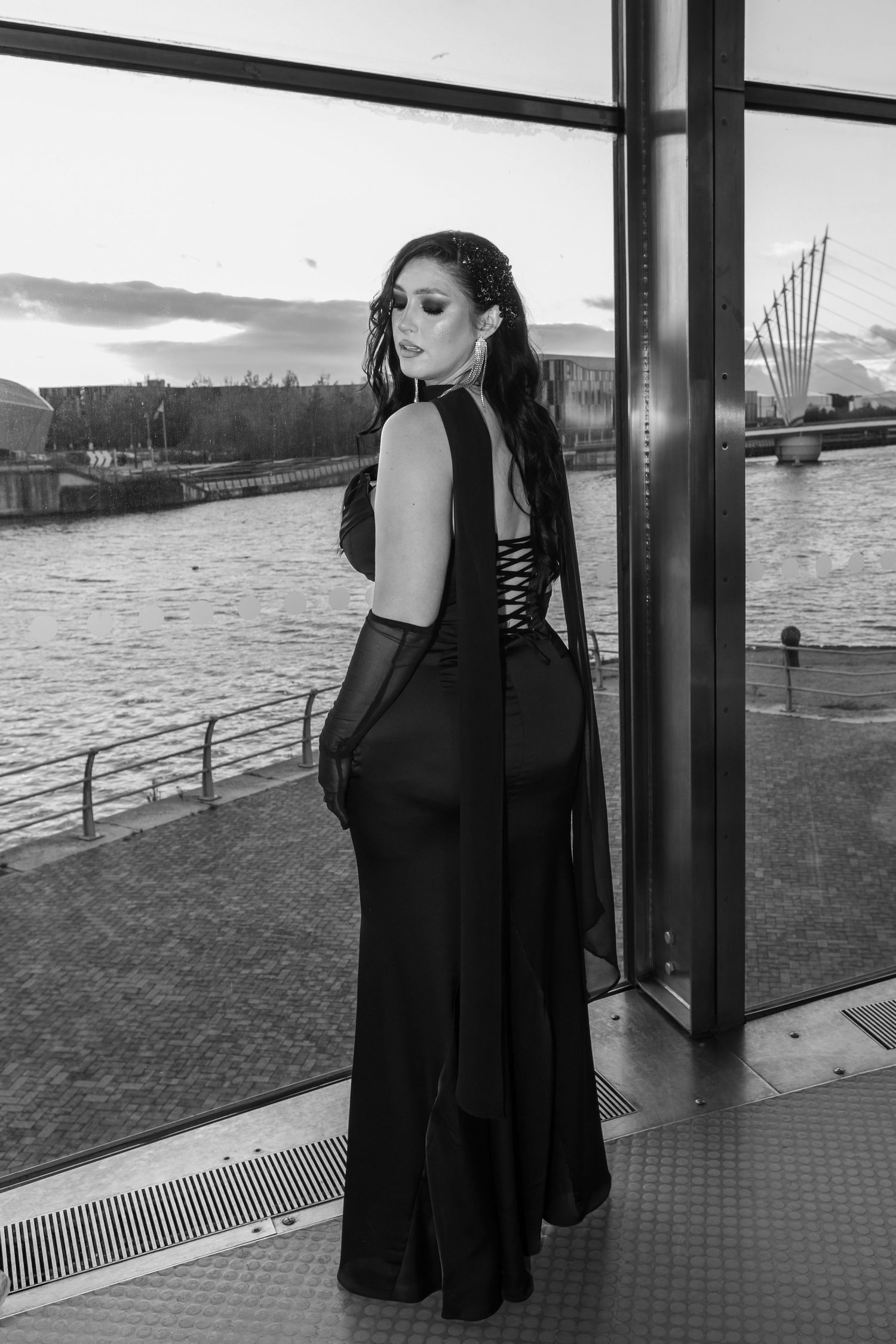 A black and white photo series showing a person in a dark dress posing near a waterfront with industrial buildings visible.