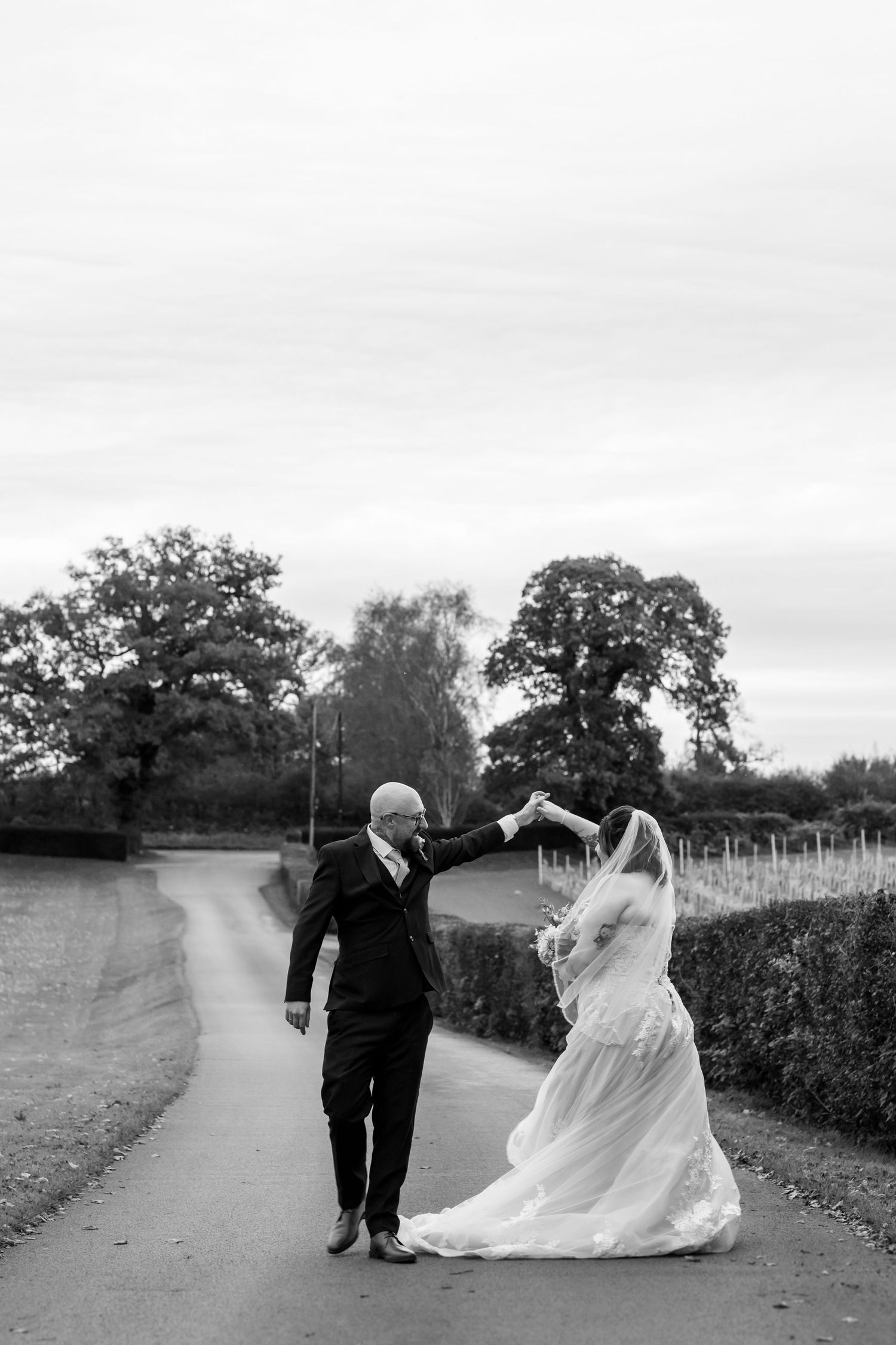 A couple walks together along a countryside path in black and white, with the bride's dress flowing in the wind.