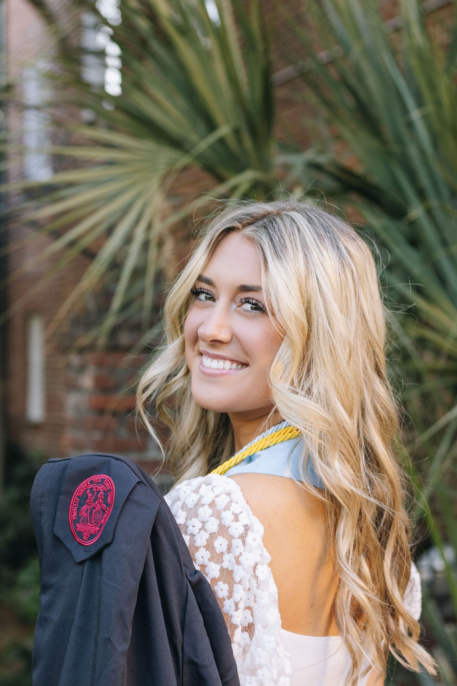 Graduate in white lace dress holds black graduation gown near palm trees.