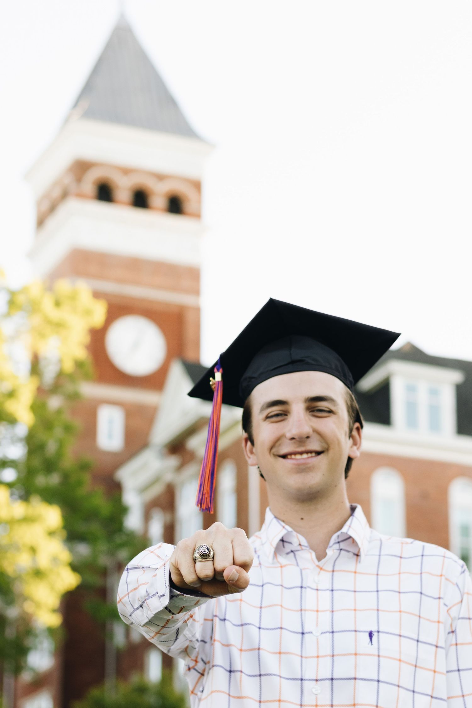 Graduate in cap points up in front of brick clock tower building.