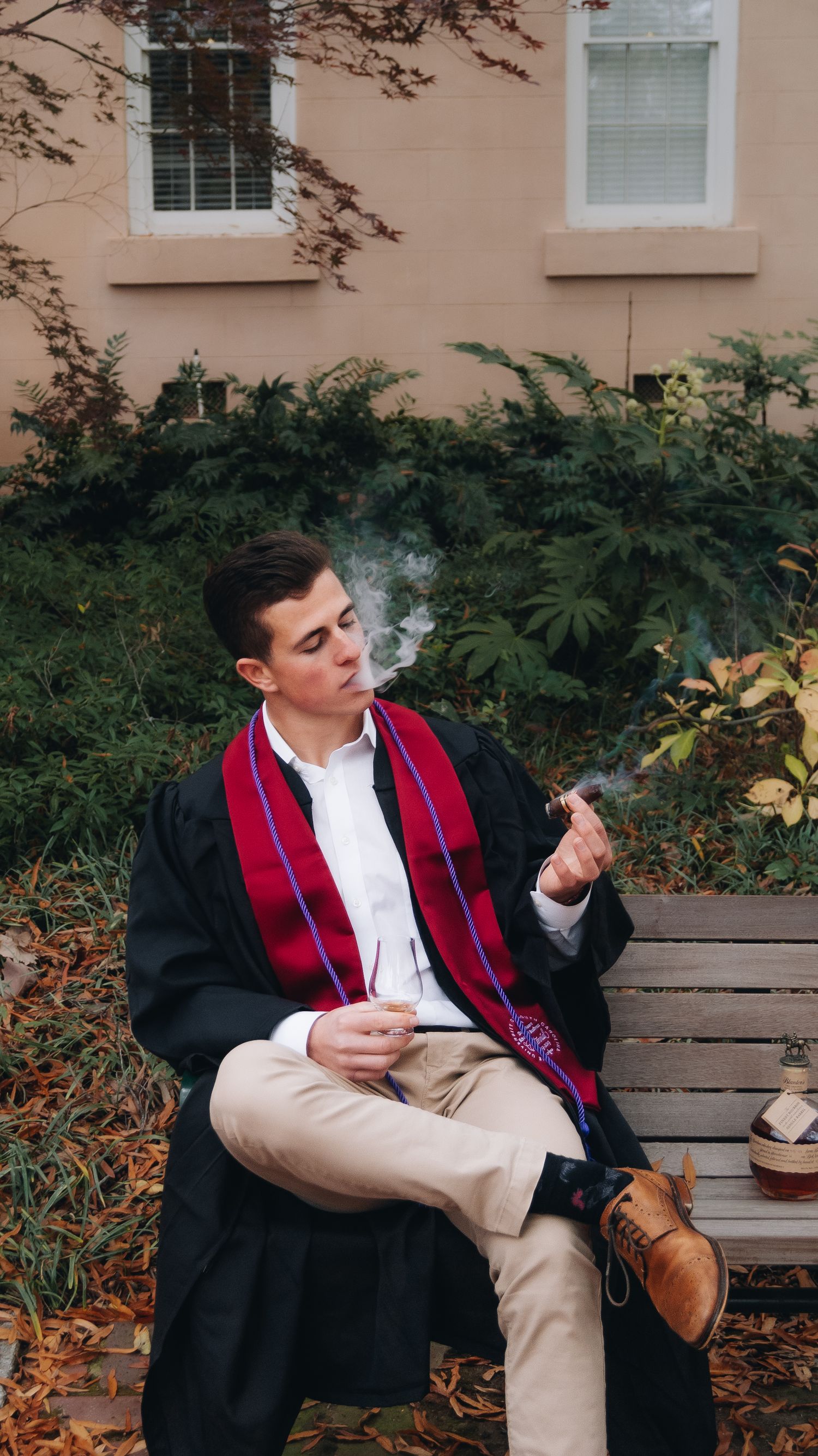 Graduate in black gown and red stole sits on bench near brick building.