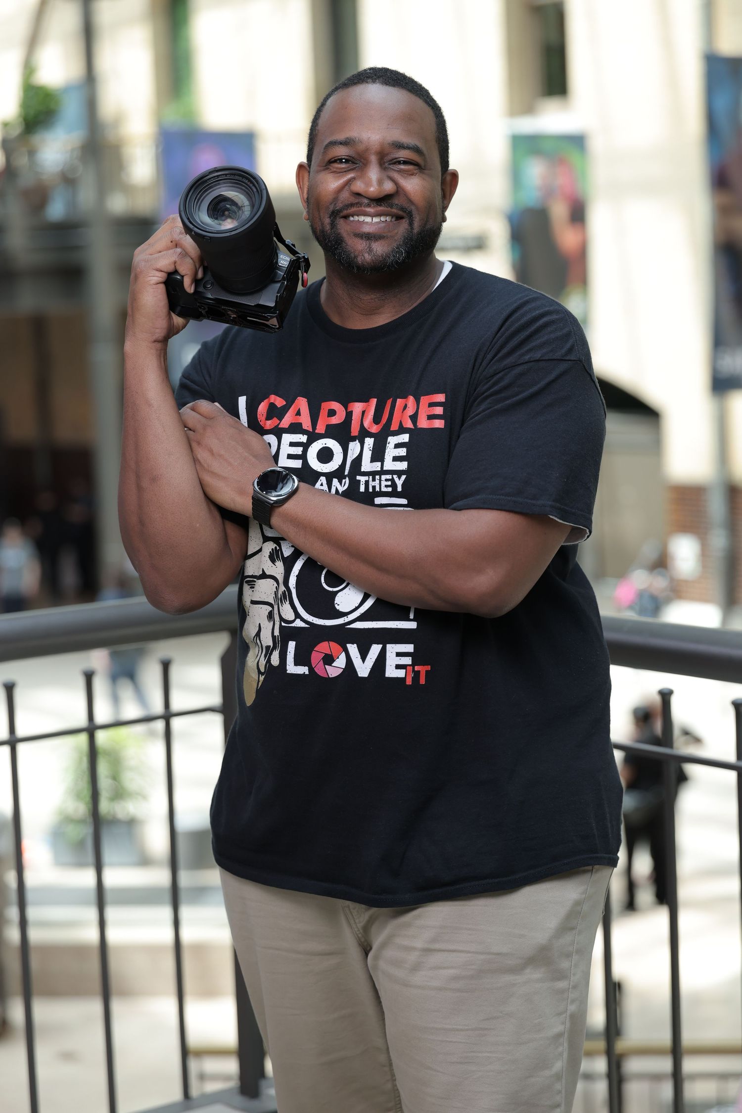 A smiling man holds a DSLR camera, wearing a black t-shirt reading I Capture People and I Love It, outdoors.