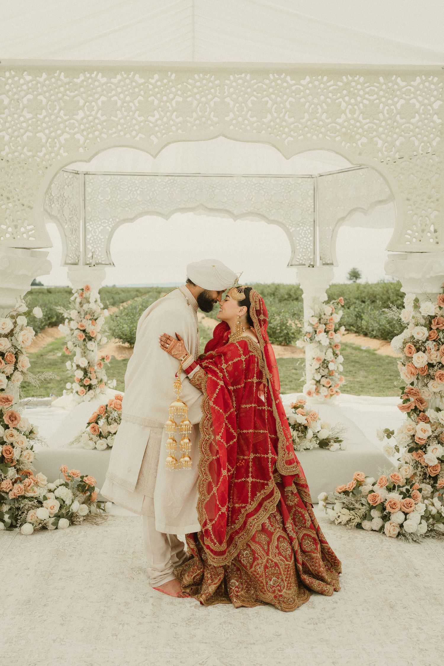 Bride in red lehenga and groom in ivory sherwani share a kiss under a floral mandap at an Indian wedding.