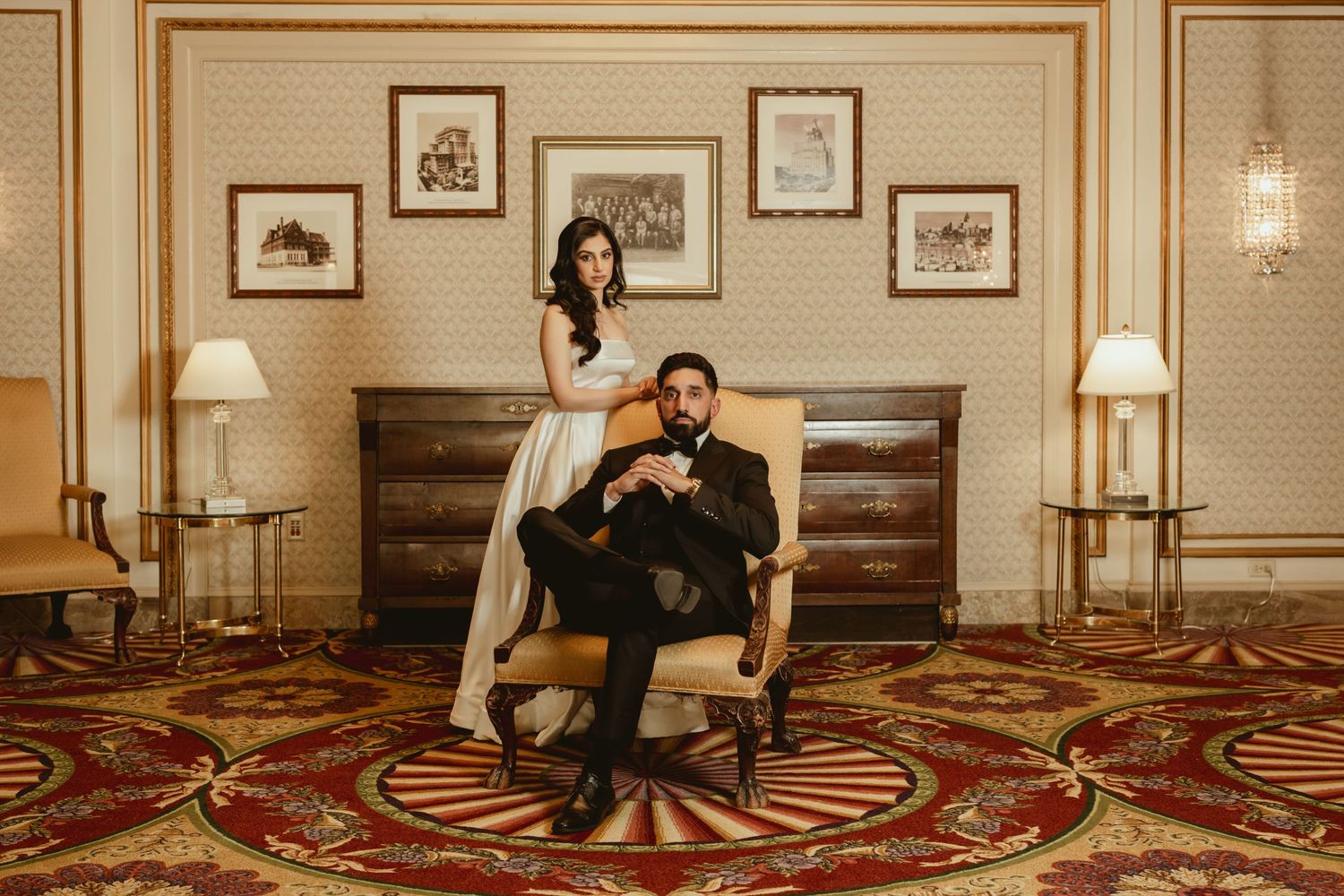 A couple poses elegantly in a vintage-style hotel room with framed artwork and ornate decor.