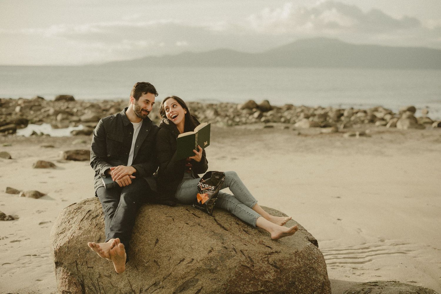 Couple relaxes on coastal rock formation while reading together at sunset.